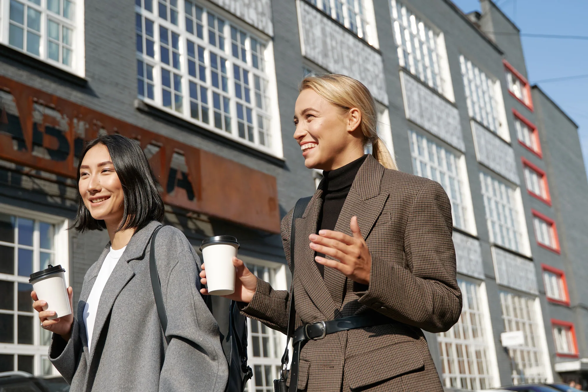 Two young women walking through the city with coffee, chatting and smiling
