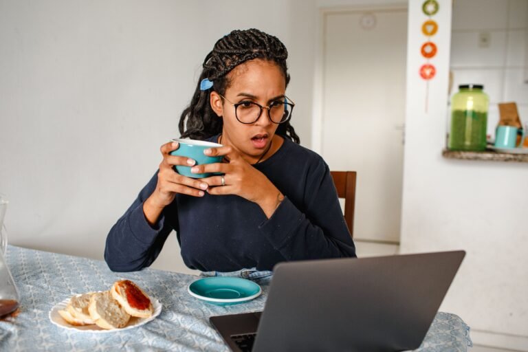 Young woman holding a coffee mug at her kitchen table with a laptop and toast nearby