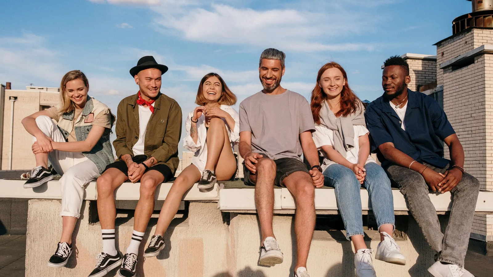 Six young adults sitting side by side on a rooftop ledge, laughing in late afternoon sunlight