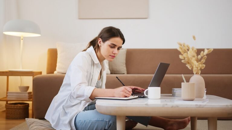 Young woman sitting on the floor at a coffee table, writing notes while working on her part-time resume on a laptop