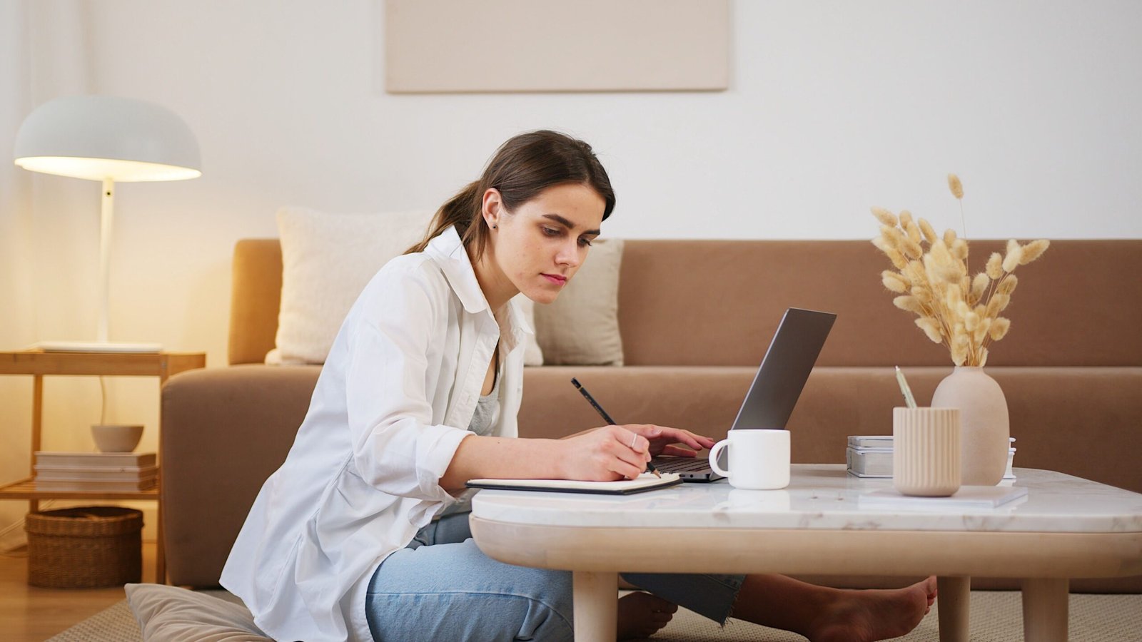 Young woman sitting on the floor at a coffee table, writing notes while working on her part-time resume on a laptop