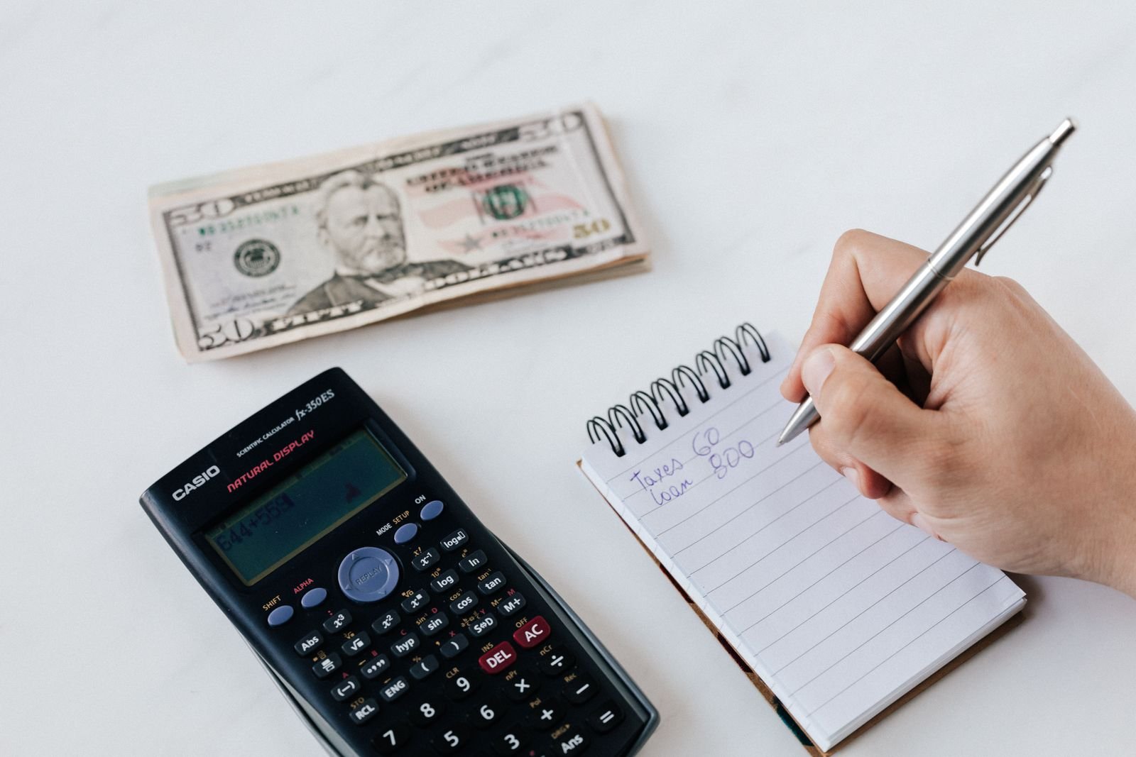 Close-up of someone writing numbers in a notebook next to a calculator and cash while planning a budget
